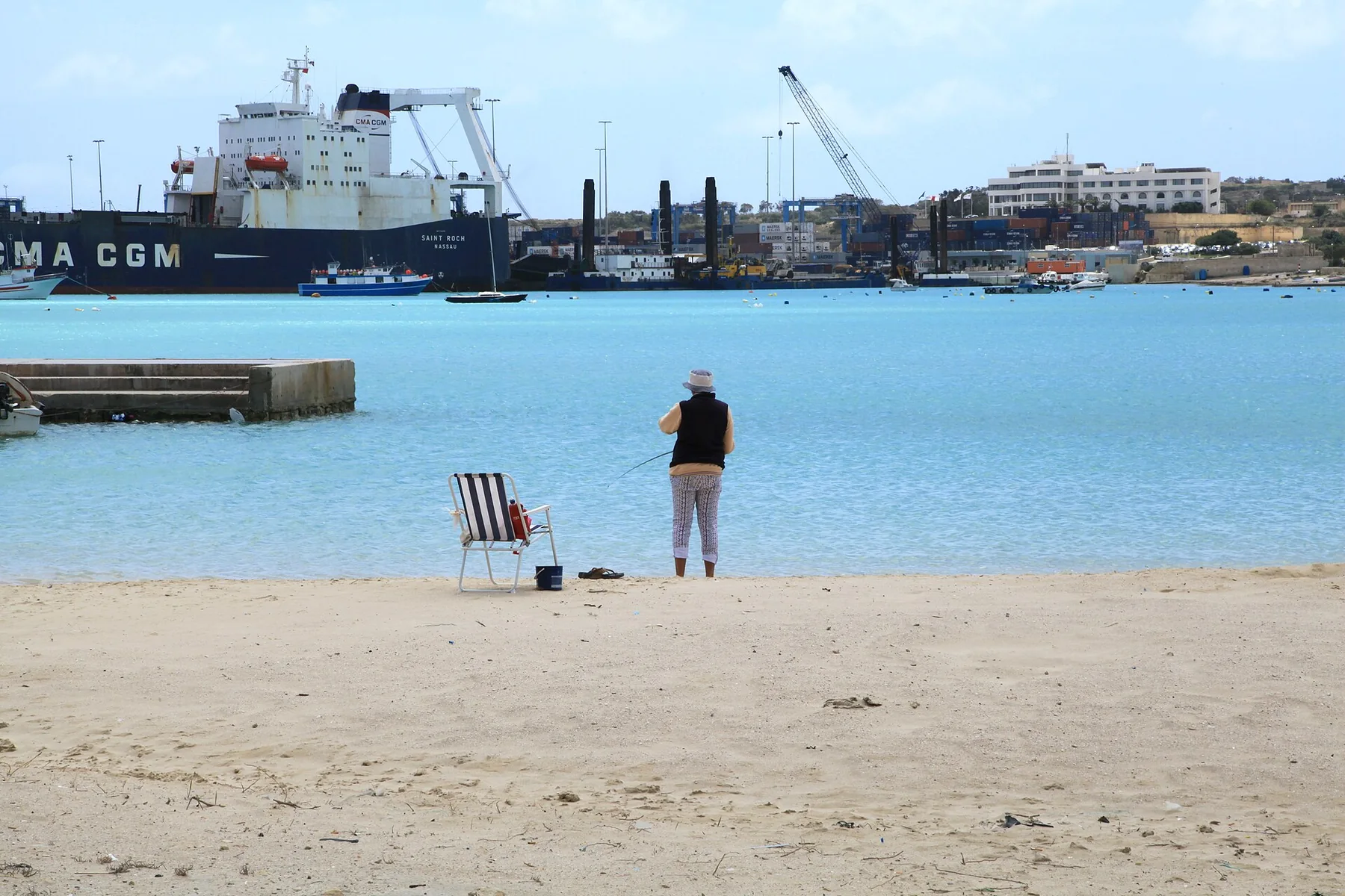 Pretty Bay in Birżebbuġa with a person fishing and port infrastructure across the water
