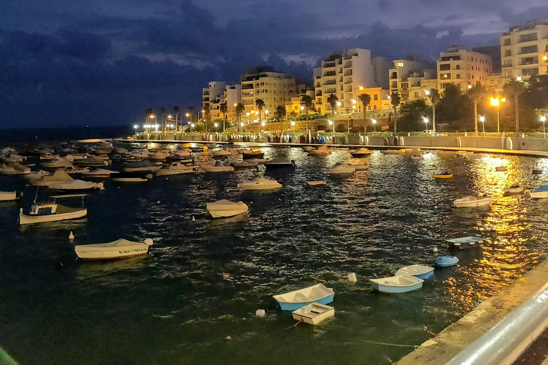 St Paul's Bay and the wider Buġibba harbour edge before sunrise, with small boats, waterfront lights and buildings